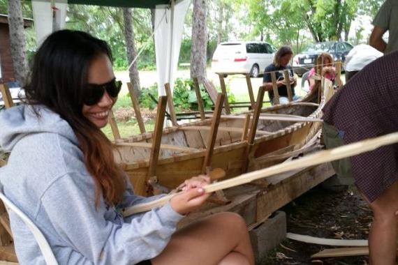 A female student holding a piece of wood beside a large wooden canoe